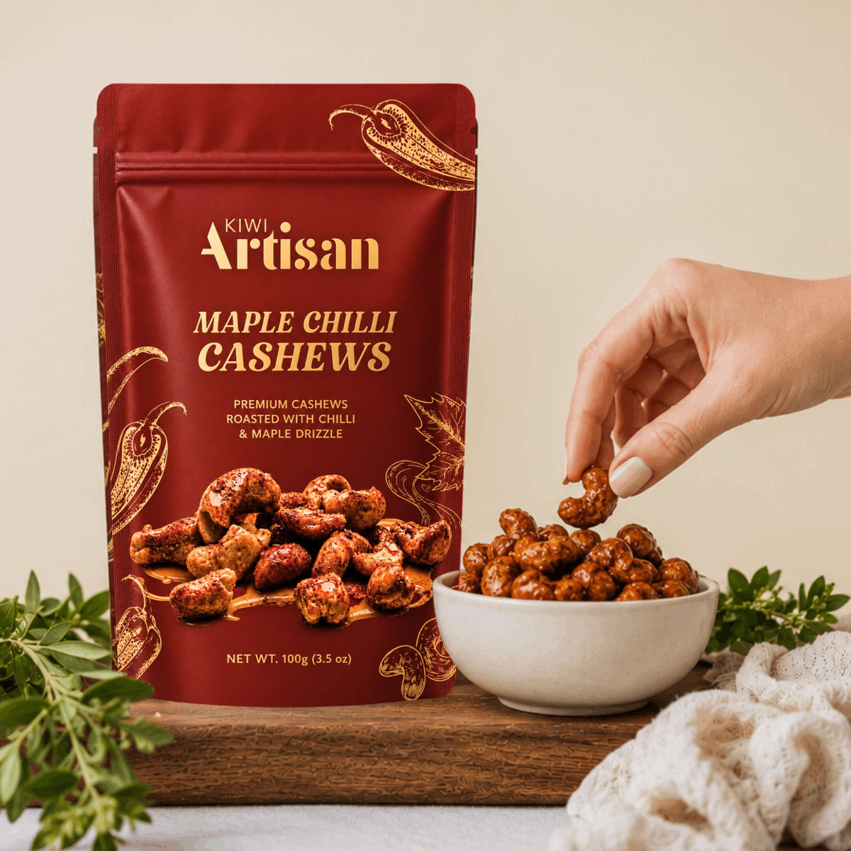 A stand-up pouch of Kiwi Artisan Maple Chilli Cashews stands on a wooden board next to a white bowl filled with the roasted cashews, with a hand reaching in to pick one. Fresh herbs and a cream knit cloth are arranged around the product against a warm beige background.
