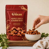A stand-up pouch of Kiwi Artisan Maple Chilli Cashews stands on a wooden board next to a white bowl filled with the roasted cashews, with a hand reaching in to pick one. Fresh herbs and a cream knit cloth are arranged around the product against a warm beige background.