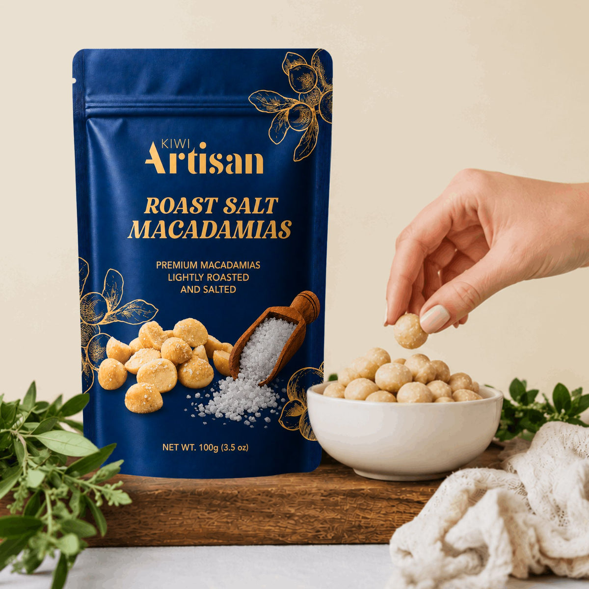 A blue stand-up pouch of Kiwi Artisan Roast Salt Macadamias stands on a wooden board next to a white bowl filled with the macadamias, with a hand reaching in to pick one. Green leaves and a beige cloth are arranged around the setup.