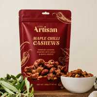 A 500g stand-up pouch of Kiwi Artisan Maple Chilli Cashews stands on a wooden surface with a small white bowl of the roasted cashews and green herbs in front. The pouch is deep red with gold lettering and decorative chili illustrations.