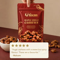 A hand holds a red stand-up pouch of Kiwi Artisan Maple Chilli Cashews against a warm beige background, with a small bowl of cashews and scattered pieces on a dark surface below. A cream review card appears at the bottom.