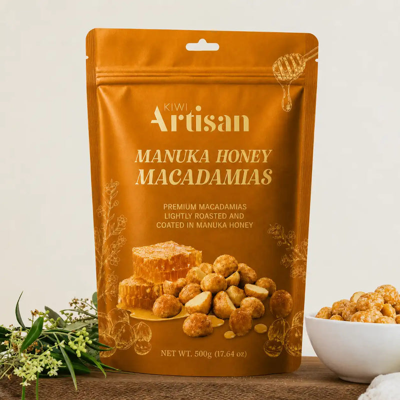 A 500g stand-up pouch of Kiwi Artisan Manuka Honey Macadamias, displayed on a wooden surface with a small white bowl of the honey-coated macadamias and greenery in the foreground.