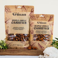 Two packages of Kiwi Artisan Maple Chilli Cashews on a wooden board with a white background.