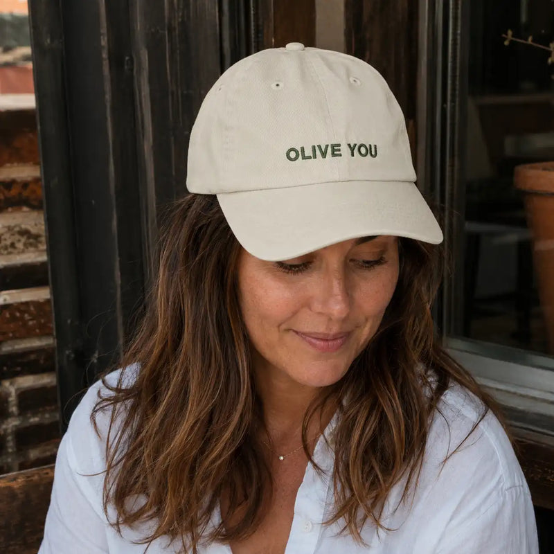 Woman wearing a beige cap with 'OLIVE YOU' text, sitting outdoors.