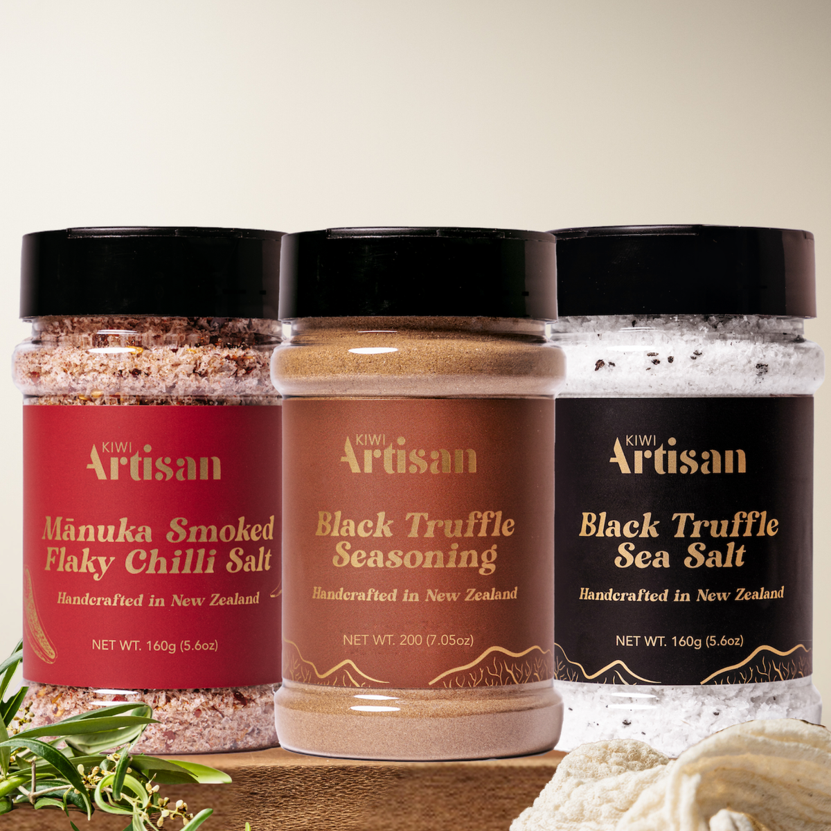 Three jars of Kiwi Artisan truffle and chili salt & seasonings on a wooden surface with a neutral background.
